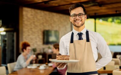 Happy waiter with slice of cake standing in a cafe and looking at camera. There are people in the background.